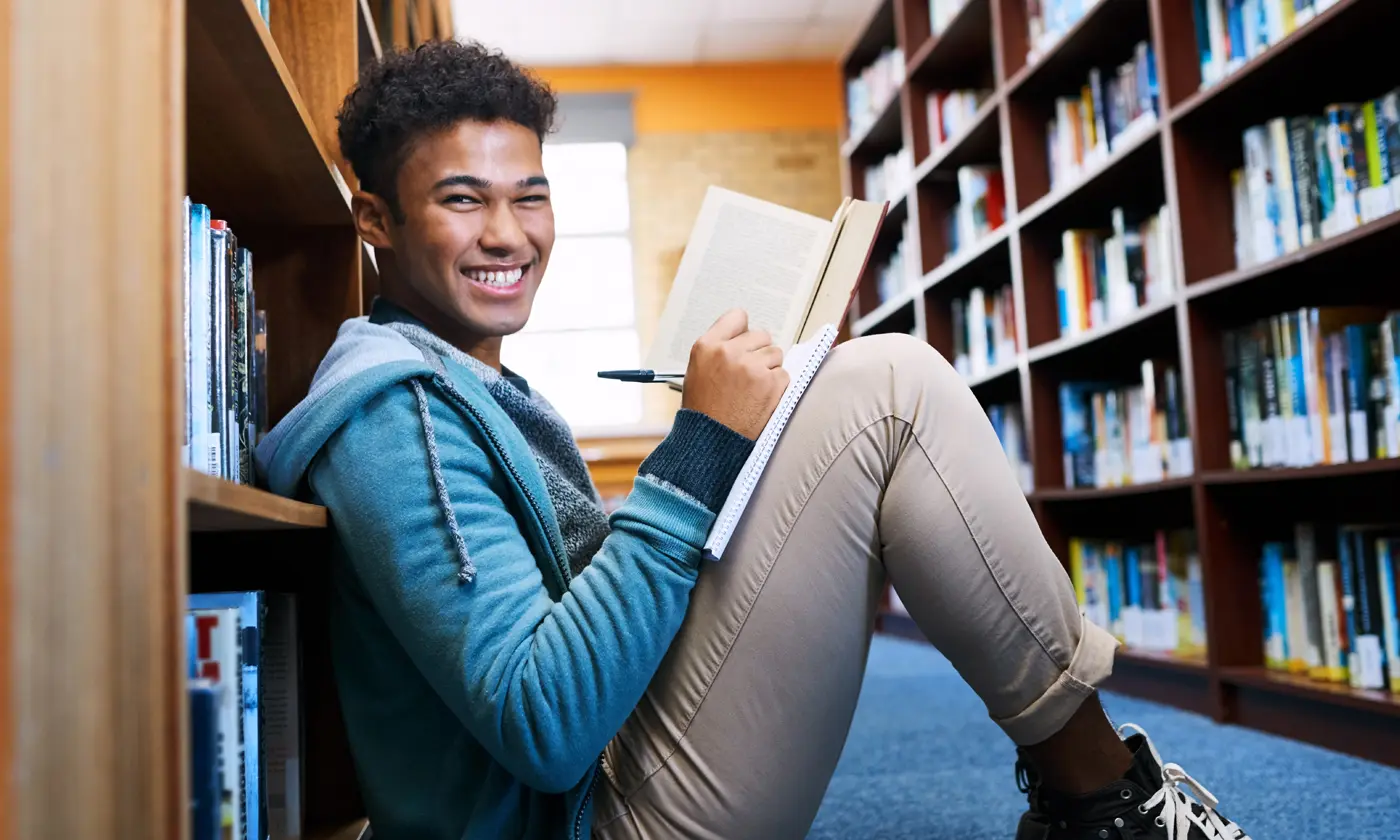 Smiling teenage boy taking notes while sitting on floor in libary